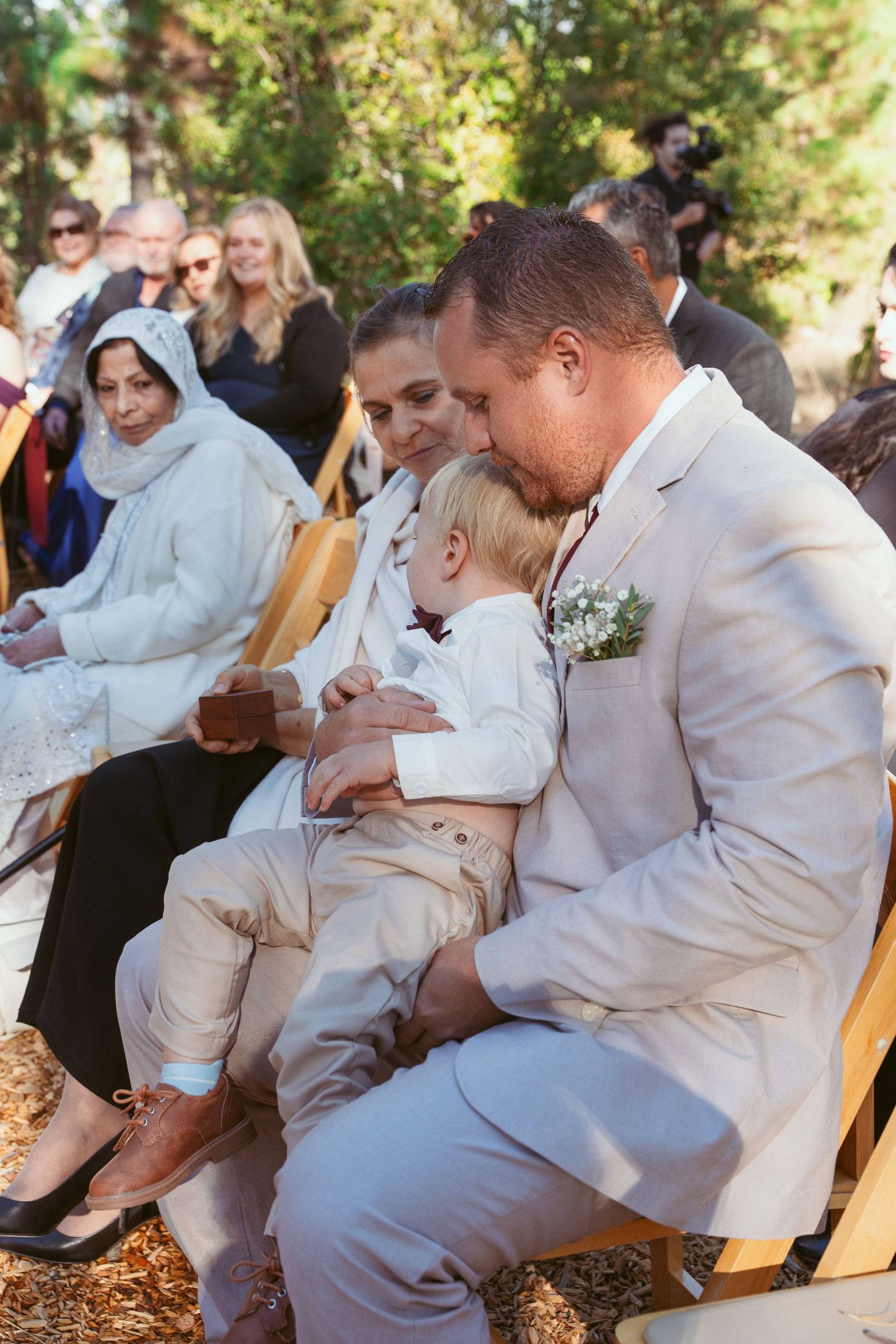 Wedding guests seated in white robes during an outdoor ceremony in a wooded setting.