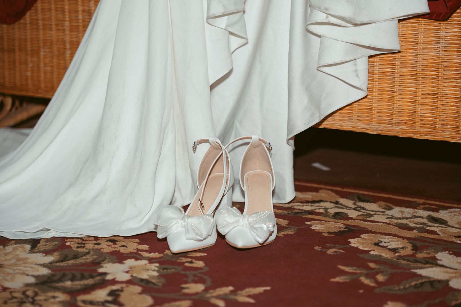 White bridal shoes and dress hem on a patterned red carpet.