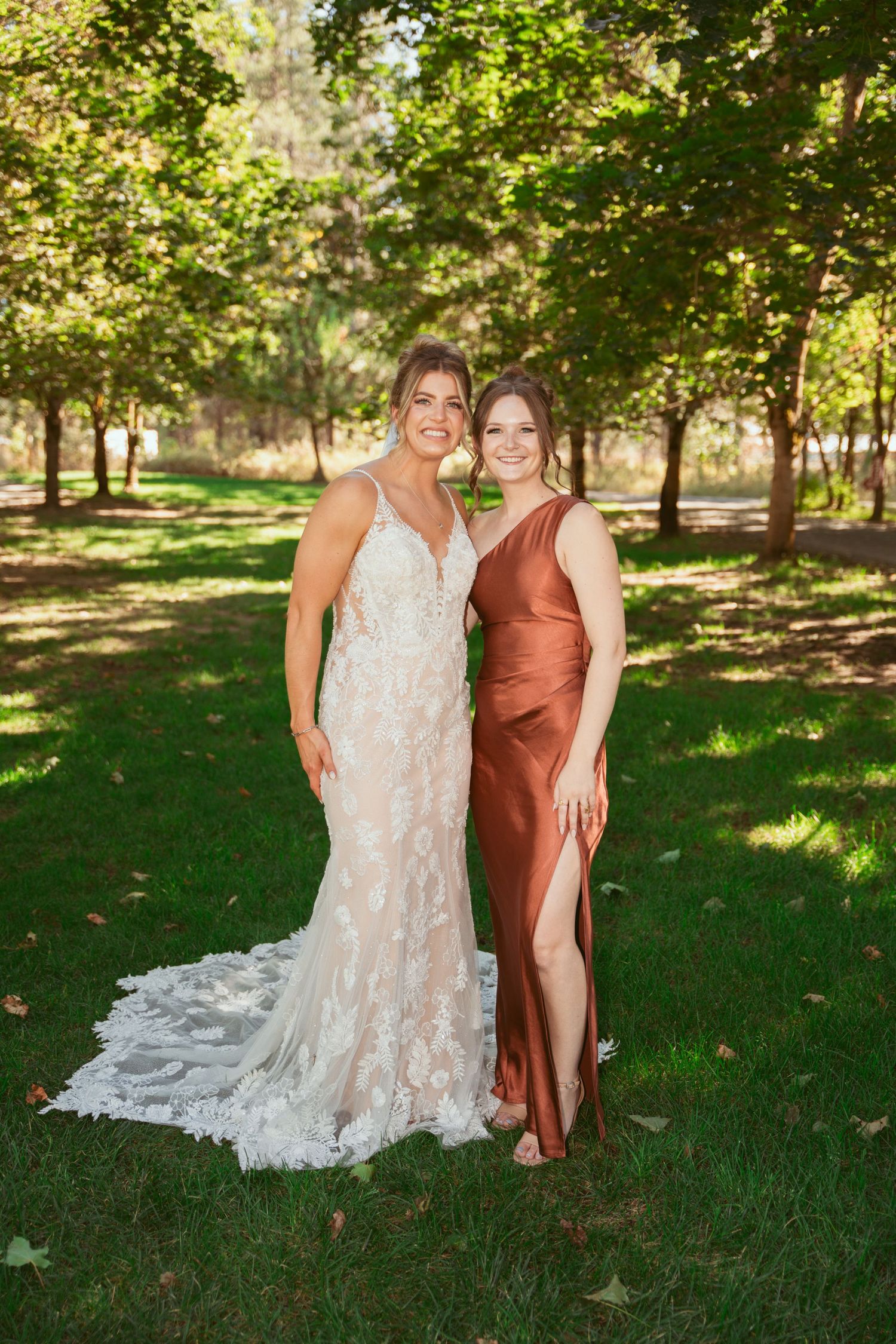 Two people in formal attire share a joyful moment outdoors, one wearing a white lace gown and the other in a copper dress.