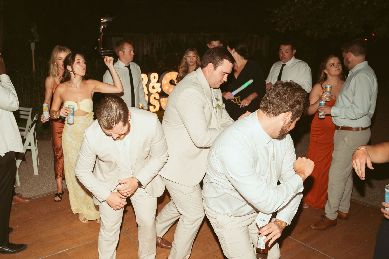 Guests in light colored suits dancing enthusiastically at an outdoor wedding reception.