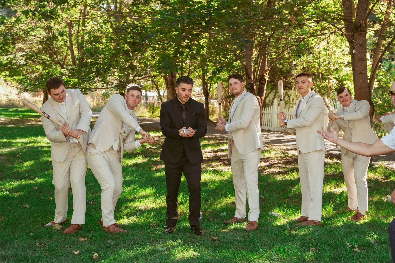 Wedding party in light colored suits poses casually on grass under trees.