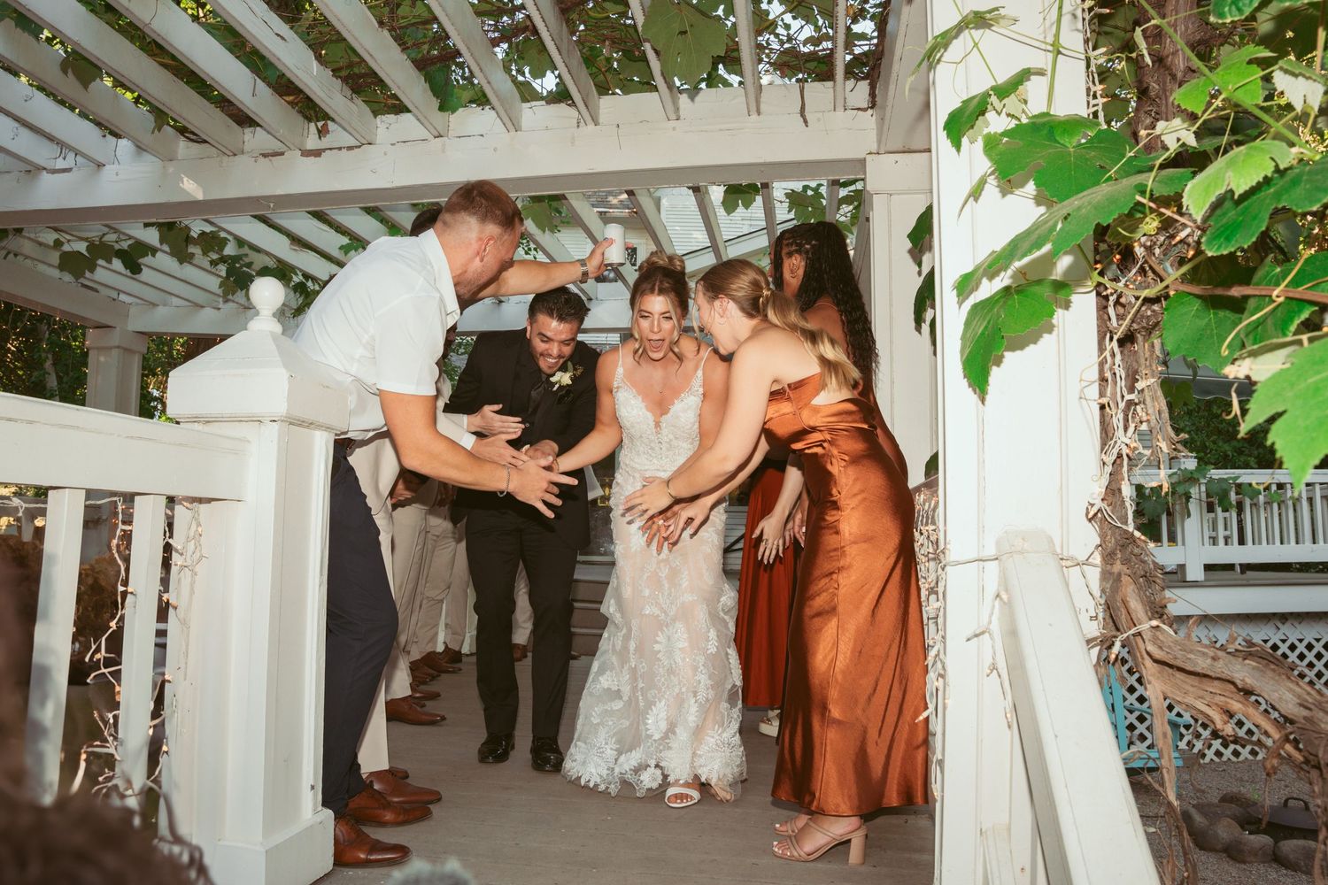 Wedding party members in rust-colored and white dresses dance on a covered outdoor patio.