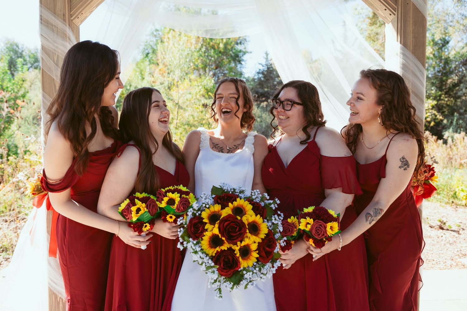 Wedding party in red dresses holding vibrant sunflower bouquets gather for photos.