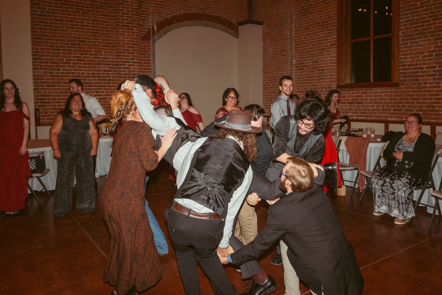 Group of party guests performing a coordinated dance move in a brick-walled reception space.