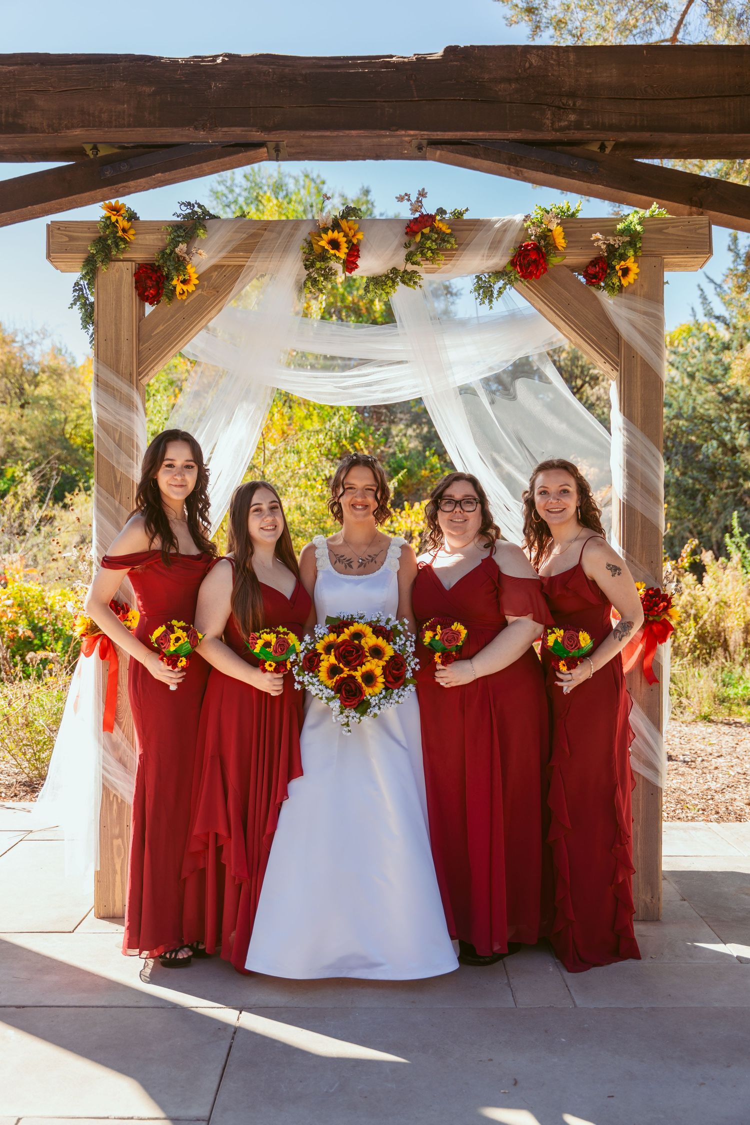 Bridal party in burgundy dresses poses under decorated outdoor pavilion with white draping.