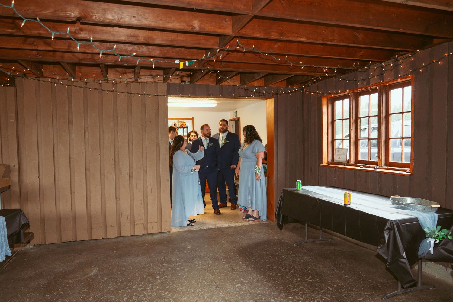 Wedding party members in light blue dresses gather near a doorway in a rustic wooden venue with string lights.