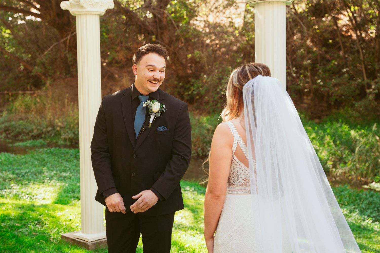 A couple in wedding attire shares a joyful moment together in a garden setting with white pillars and greenery.