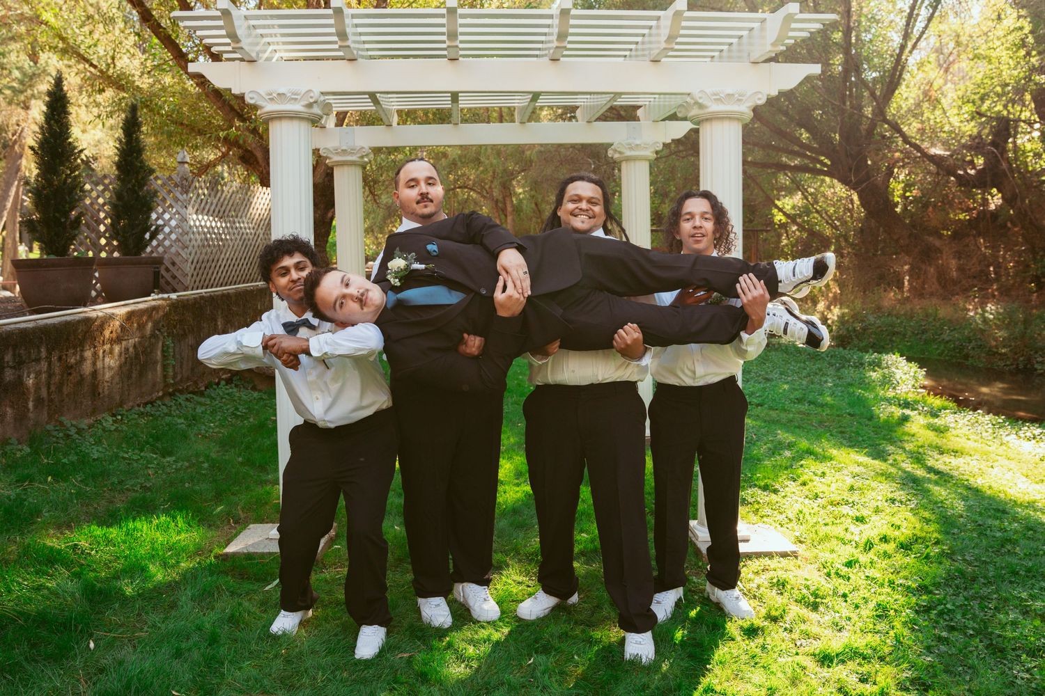 Groomsmen having fun with playful group lift pose under white garden pergola.