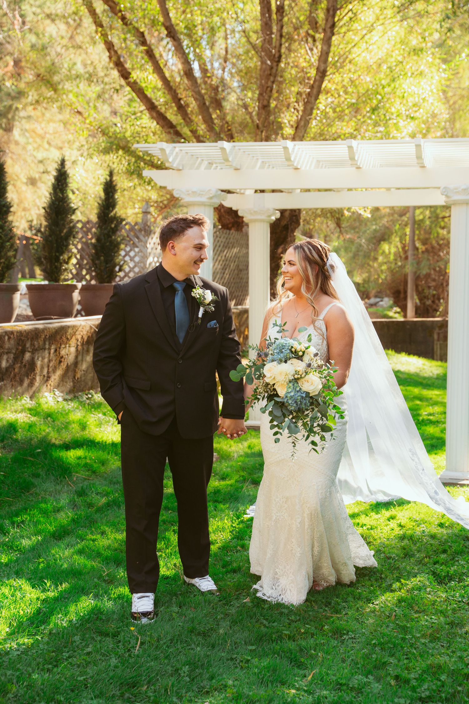 A wedding couple poses for photos on a sunny day under a white pergola surrounded by green grass.