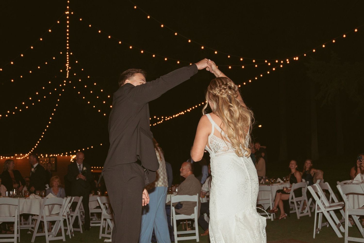 Wedding guests dance under twinkling string lights at an outdoor evening reception.