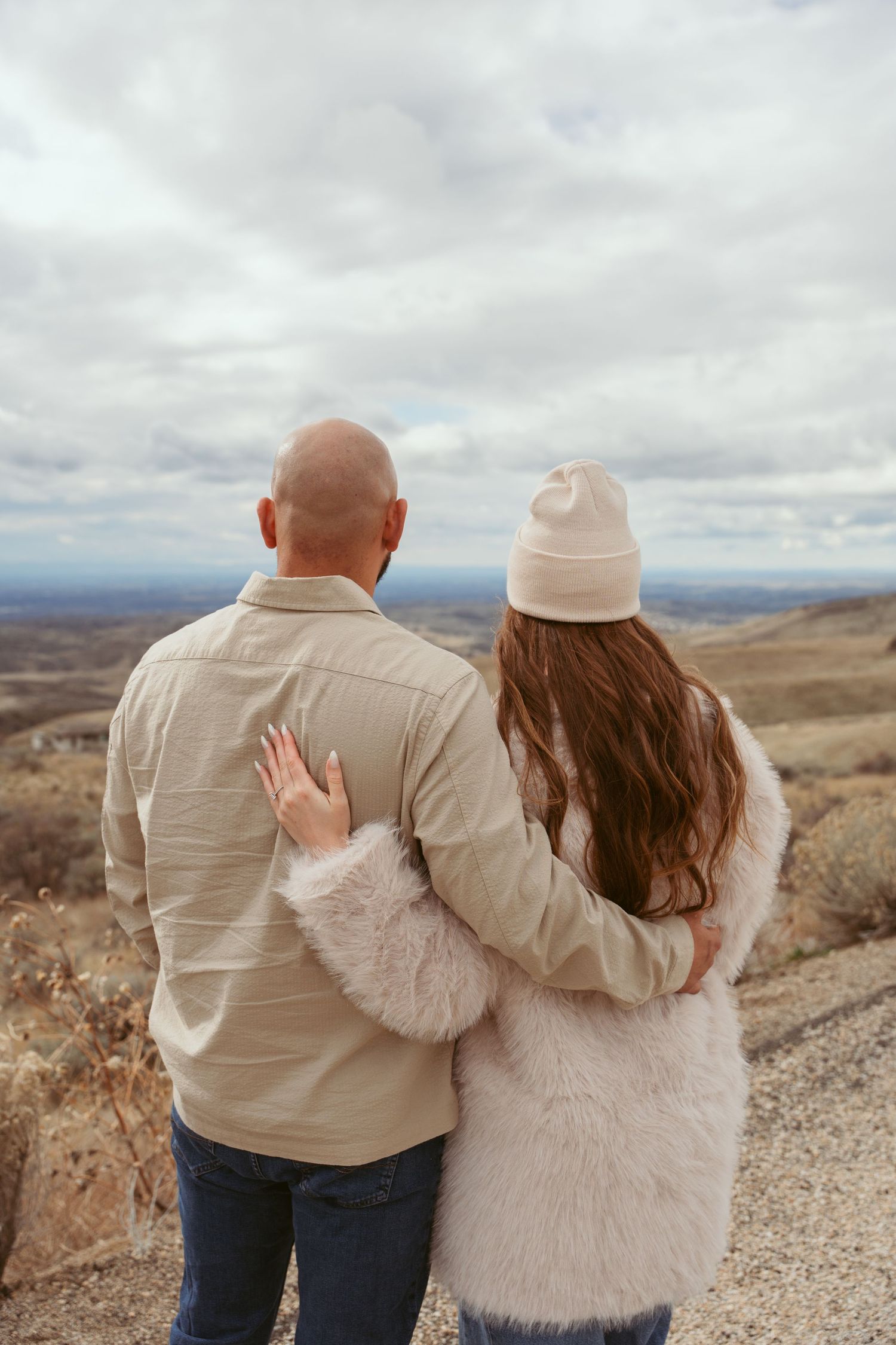 A couple stands together overlooking a vast desert landscape under cloudy skies.