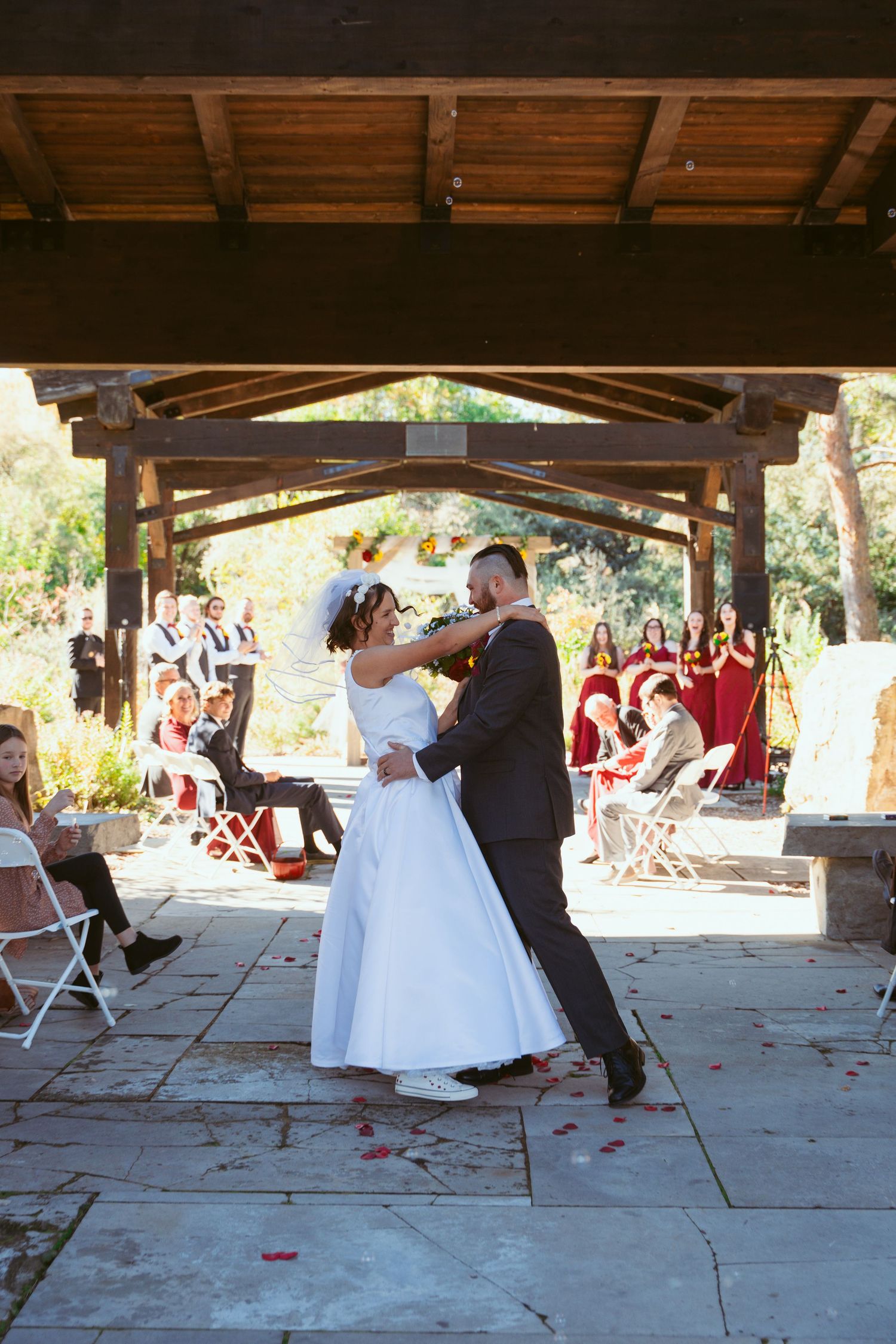 A wedding reception under a rustic wooden pavilion with guests seated at tables and white folding chairs.