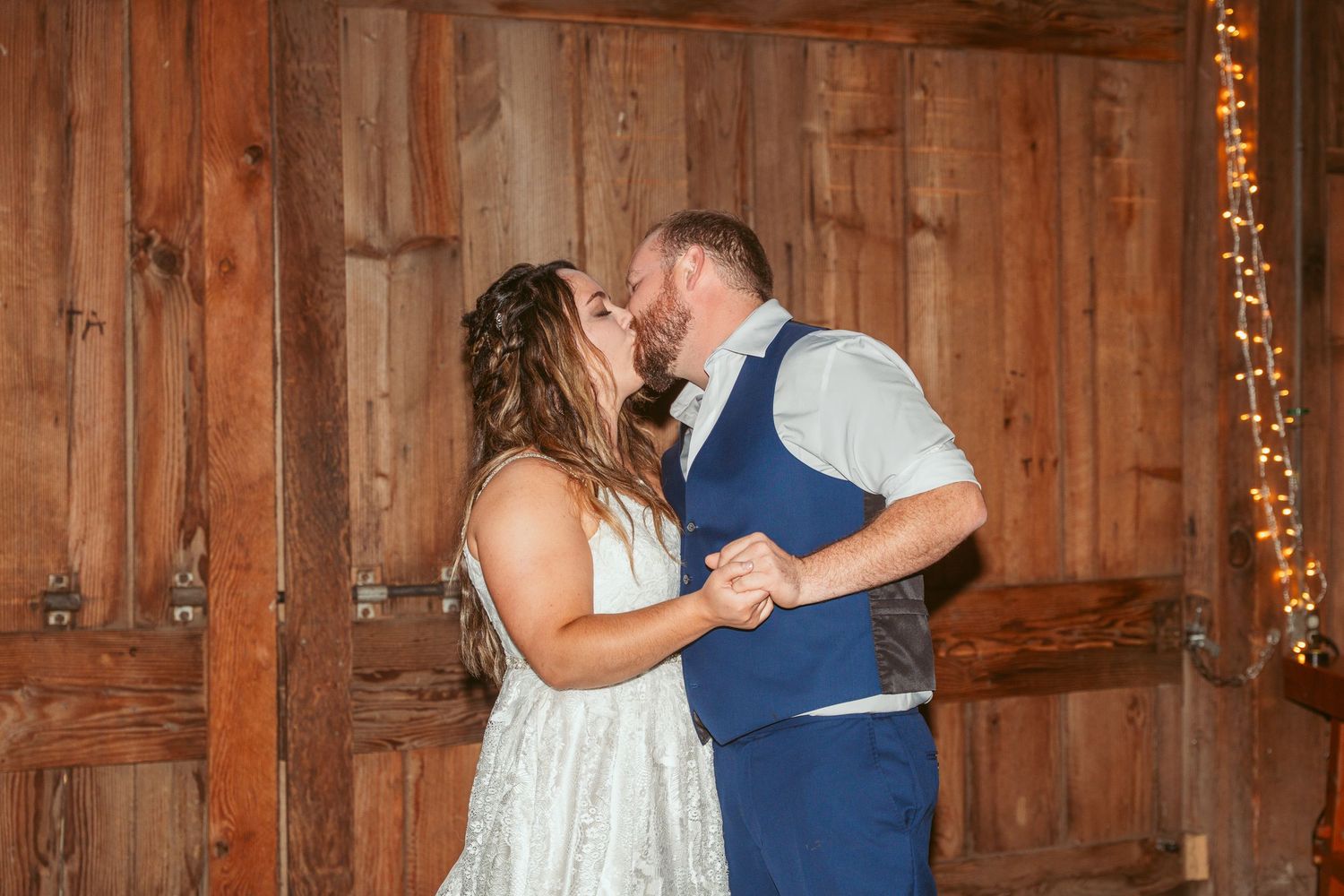 A couple shares their first dance in a rustic barn venue decorated with string lights.