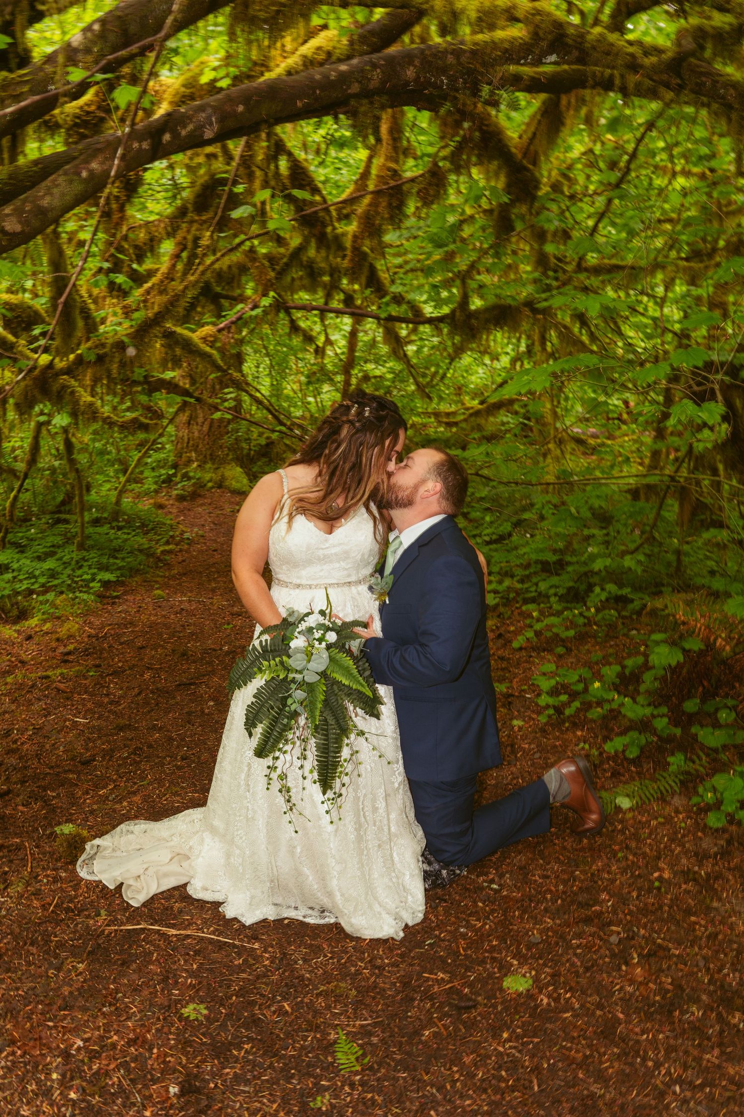 Newlyweds share intimate moments under mossy forest canopy.