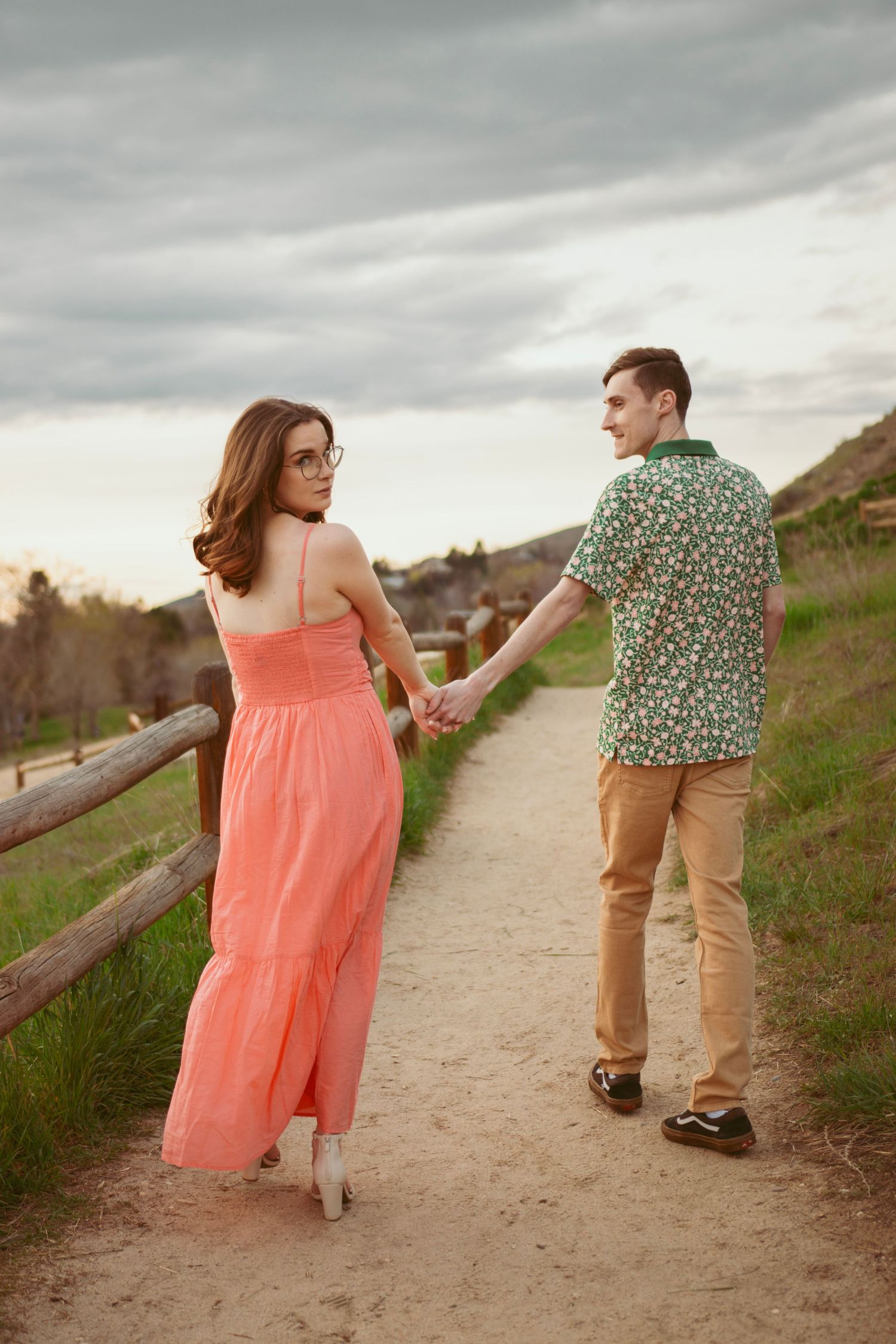 A couple in coral dress and floral shirt hold hands while walking on a hillside path.