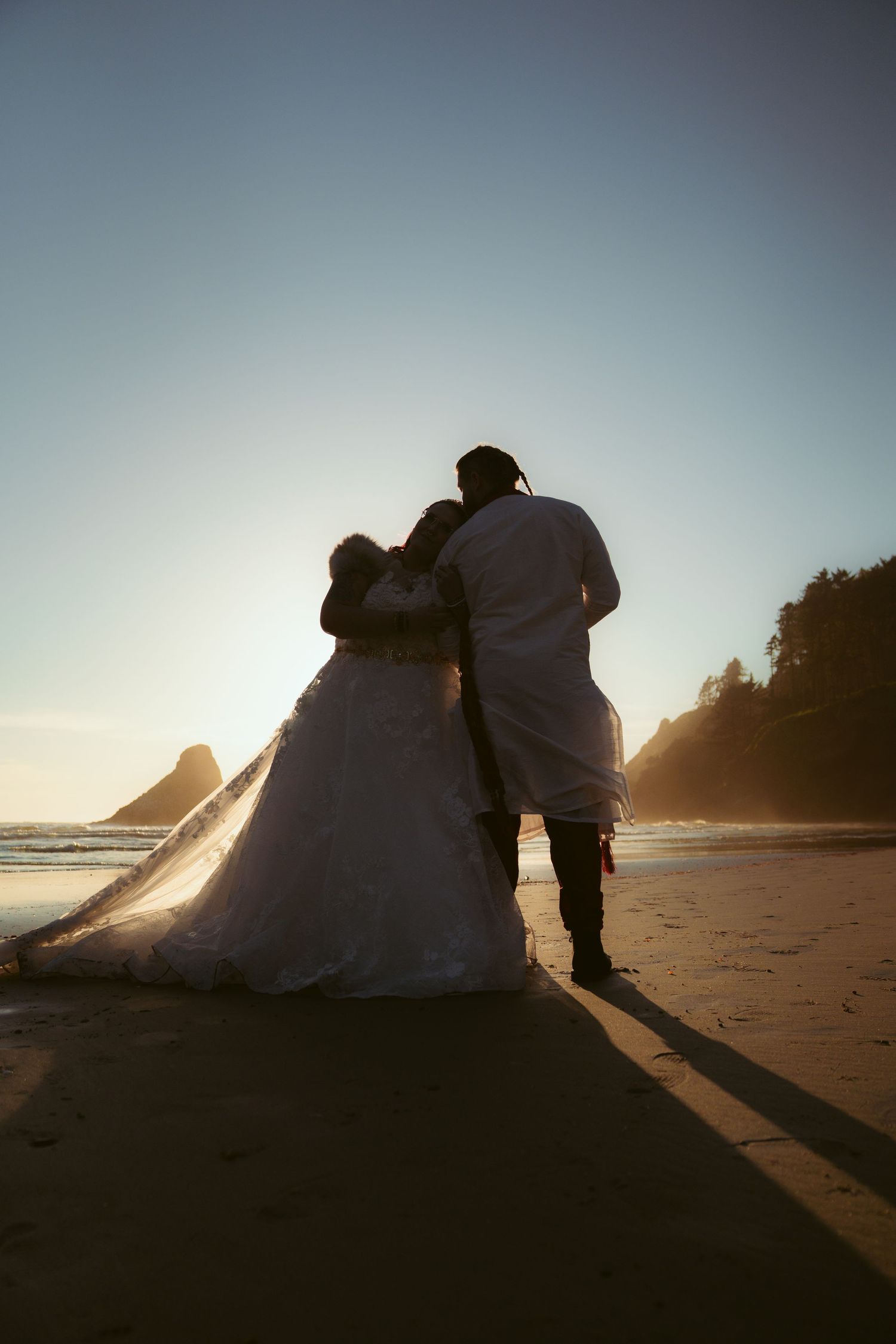 Silhouette of newlyweds walking along a beach at sunset with coastal cliffs in view.