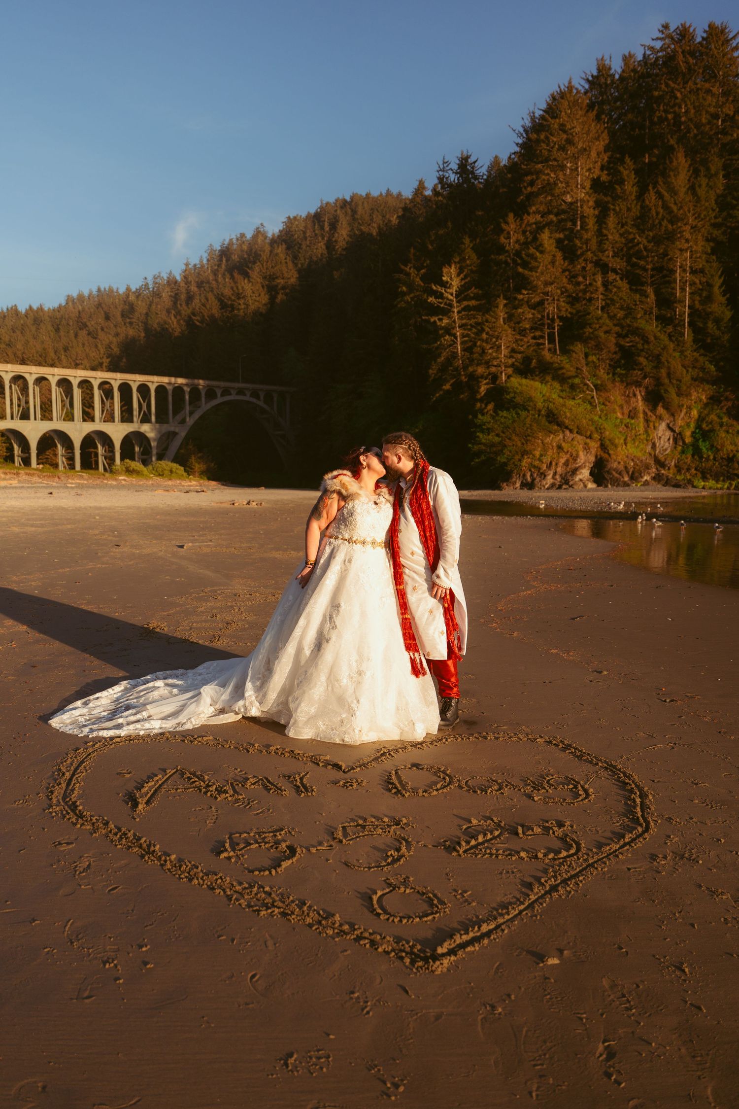 Wedding couple posing on sandy beach with heart drawn in sand at sunset.