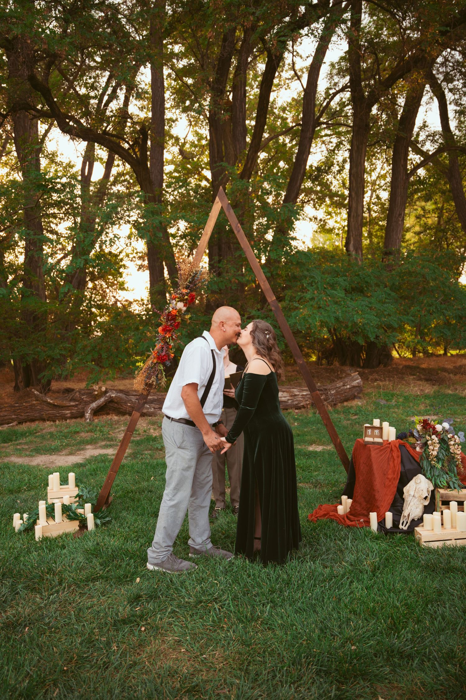 A couple stands beneath a triangle arch with romantic candle decorations in a wooded setting.