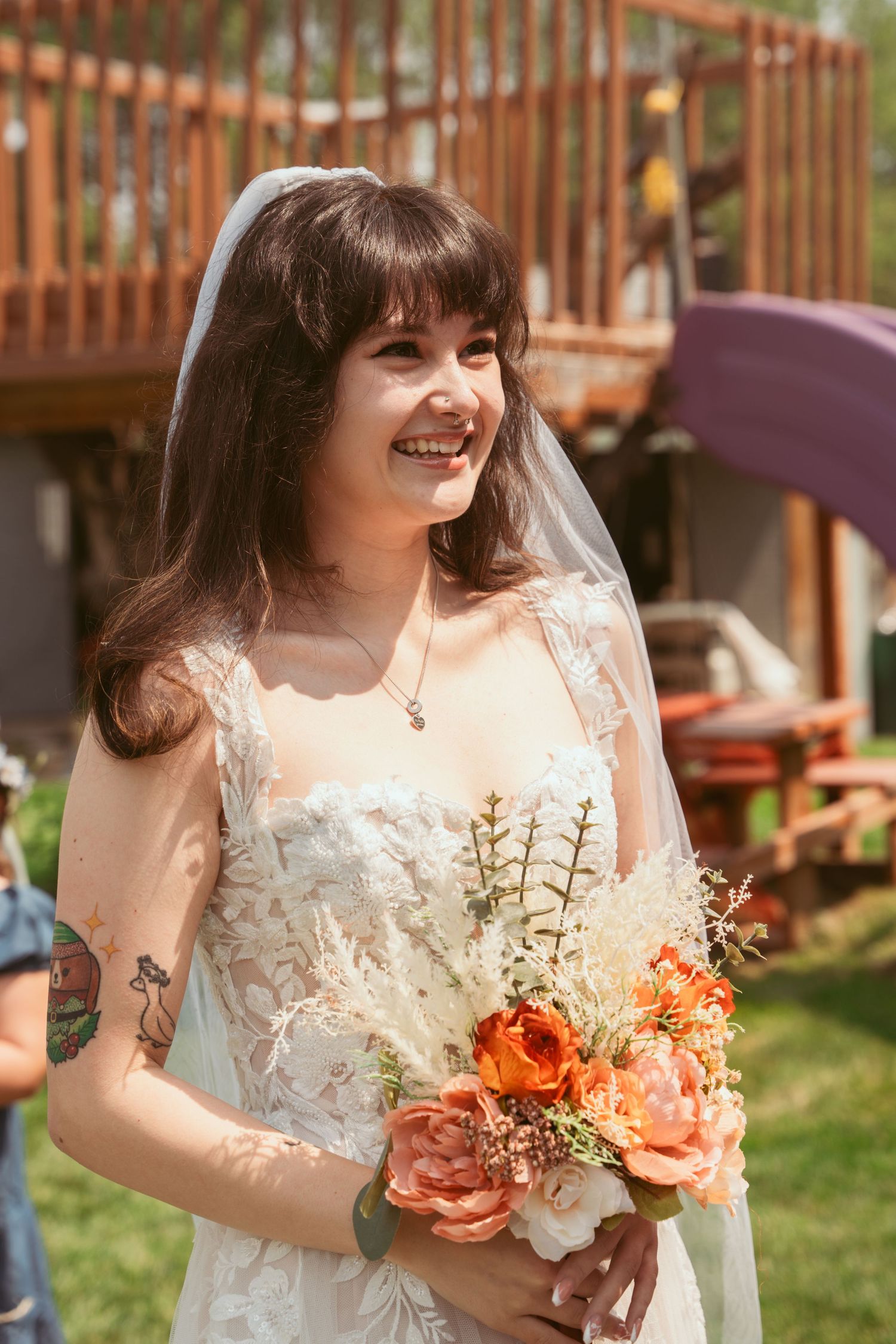 Bride in white lace dress holds coral-colored bouquet in outdoor garden setting.