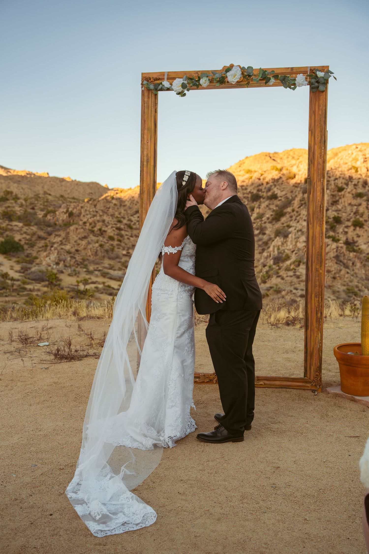 A couple embraces during their wedding ceremony in front of a wooden arch in a desert setting.