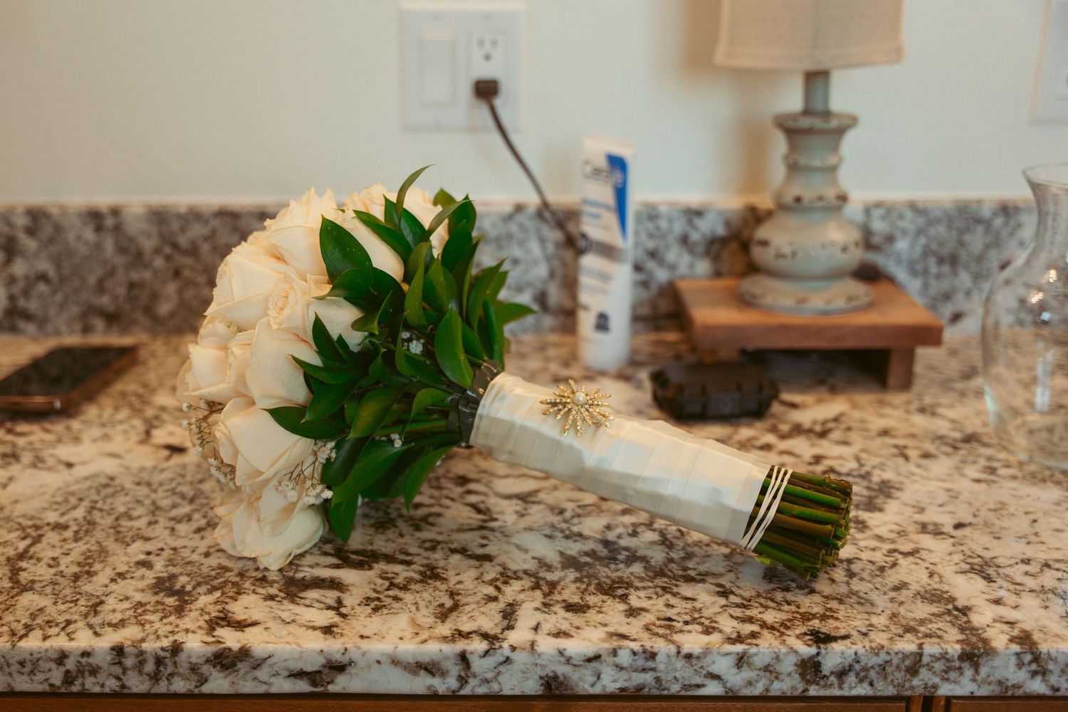 Green plant leaves next to a wooden ring box on a speckled marble counter surface.