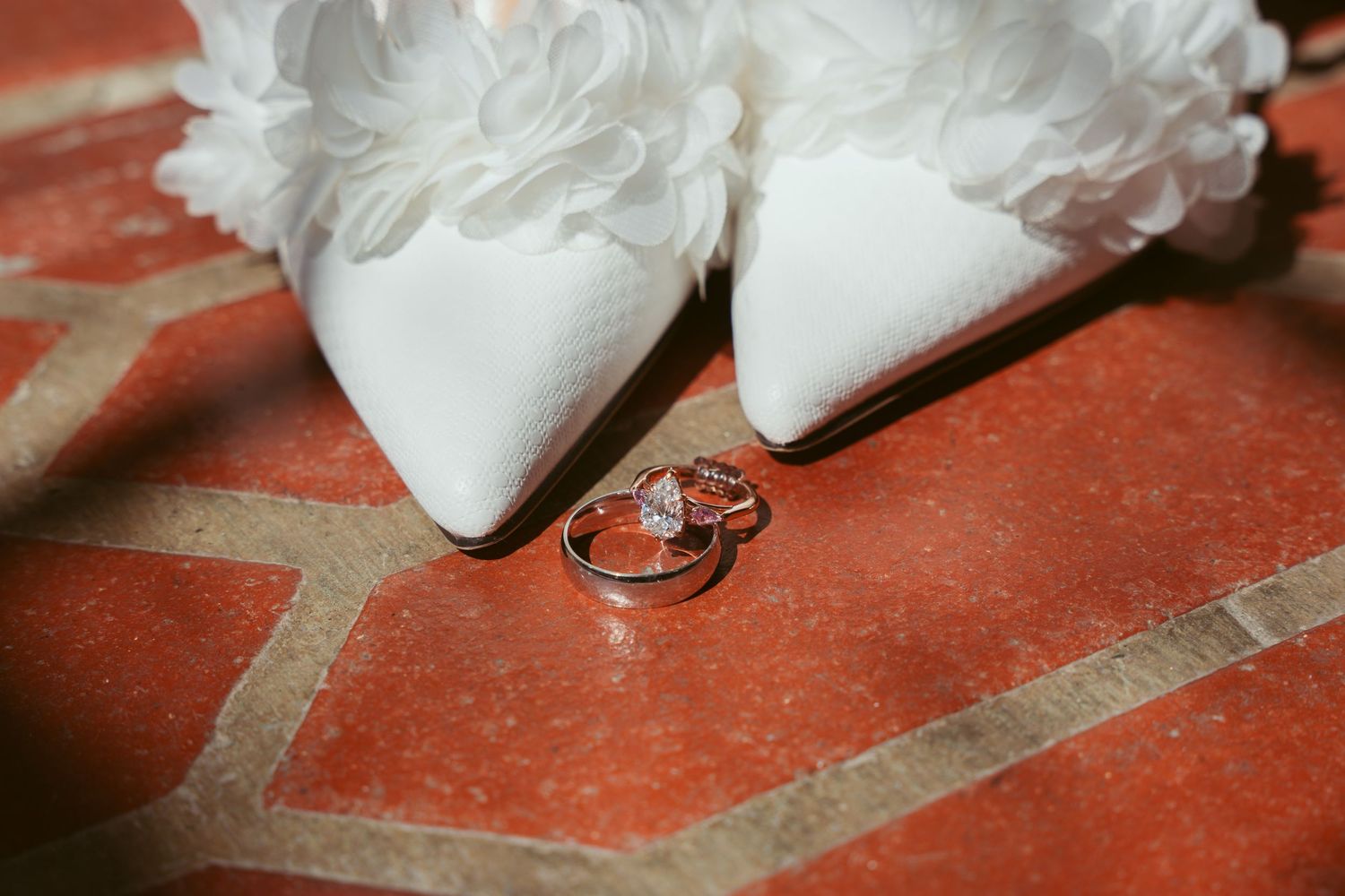 Wedding rings placed in front of white bridal shoes on a terracotta tile floor.