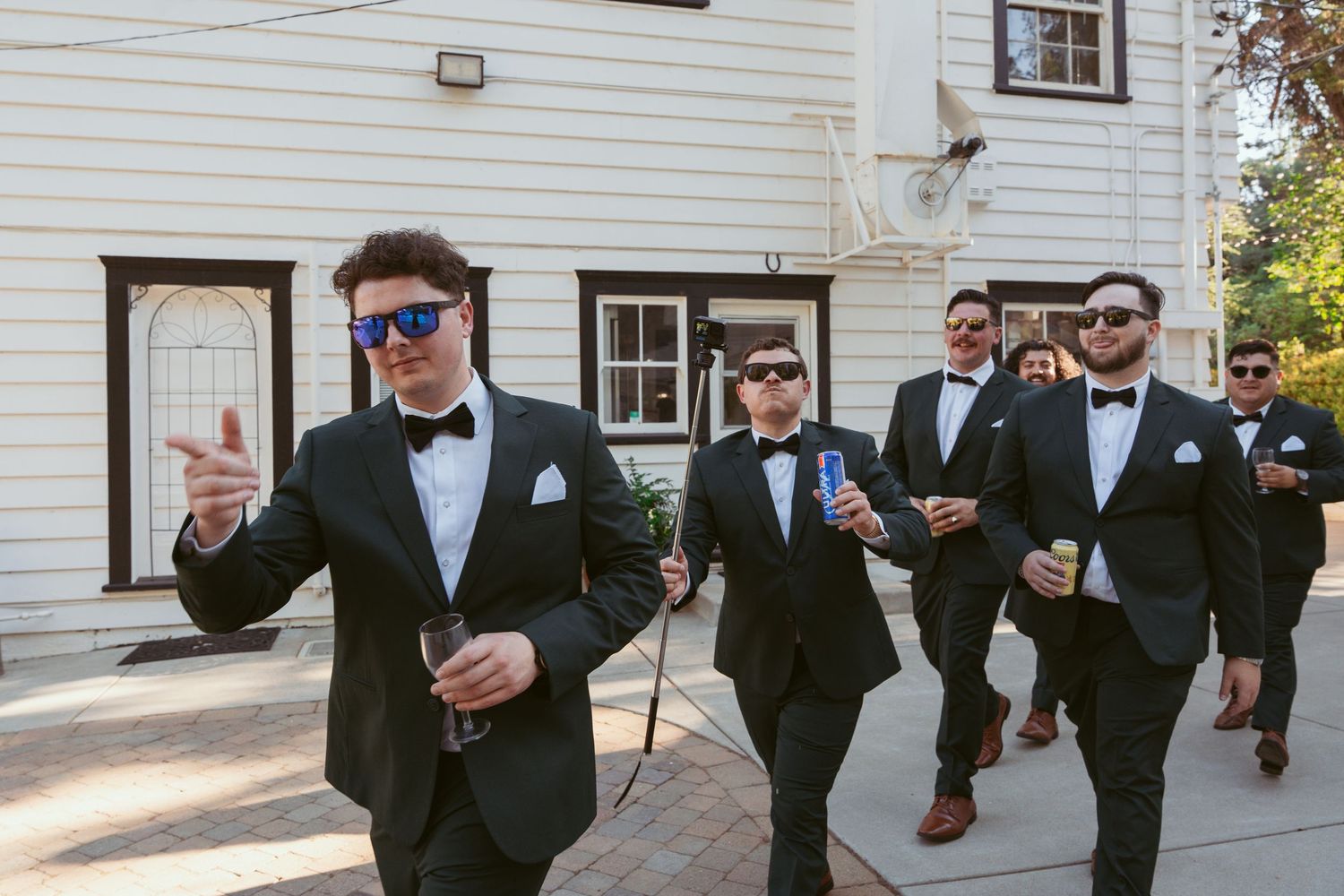 Groomsmen in black tuxedos walk together outside a white building during wedding festivities.