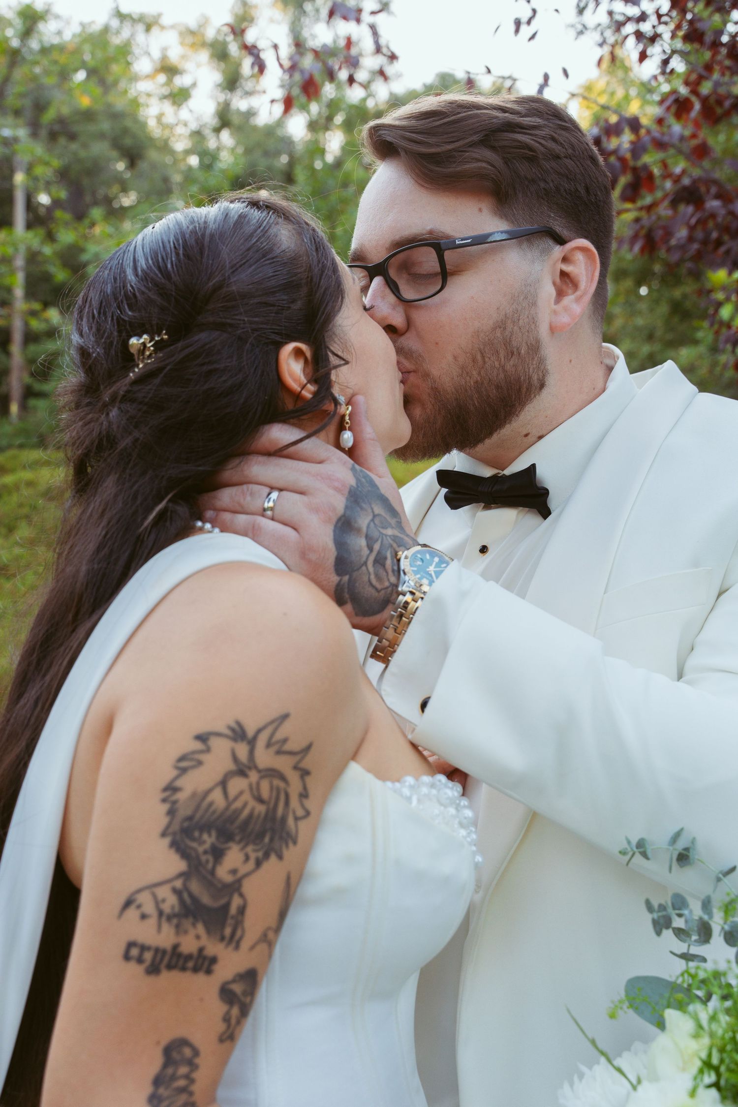 A close-up of a wedding couple sharing a kiss outdoors with visible tattoo art.