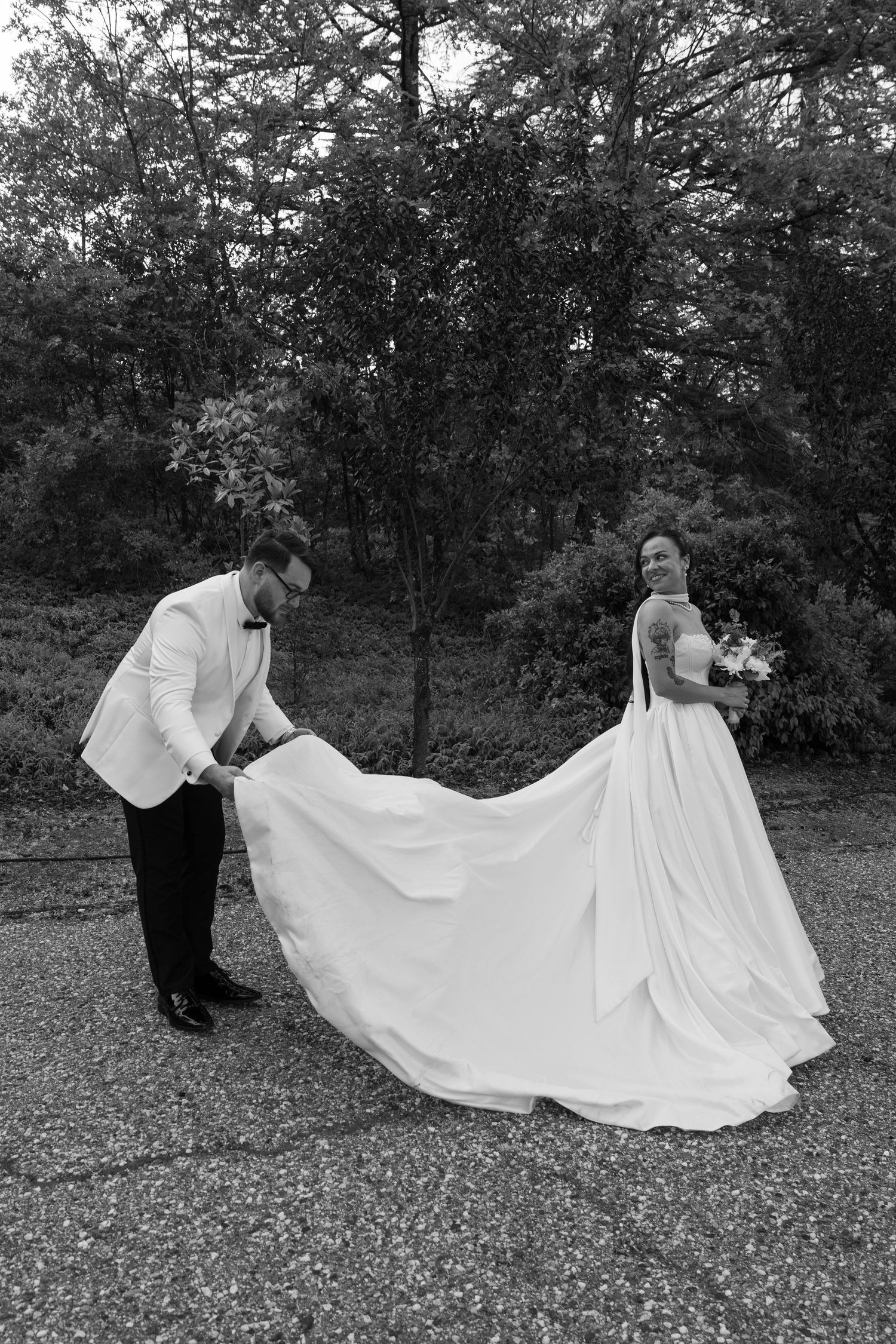 A black and white wedding photo showing the train of an elegant white dress being arranged outdoors.