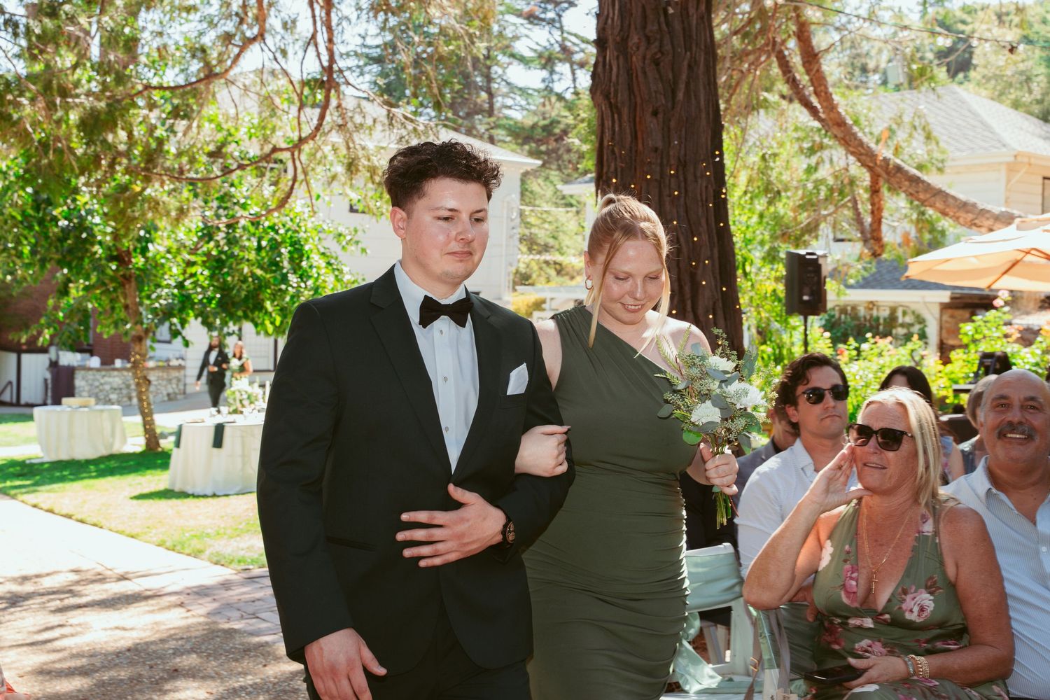 A group of wedding guests gather outdoors under tall trees during a sunny daytime celebration.