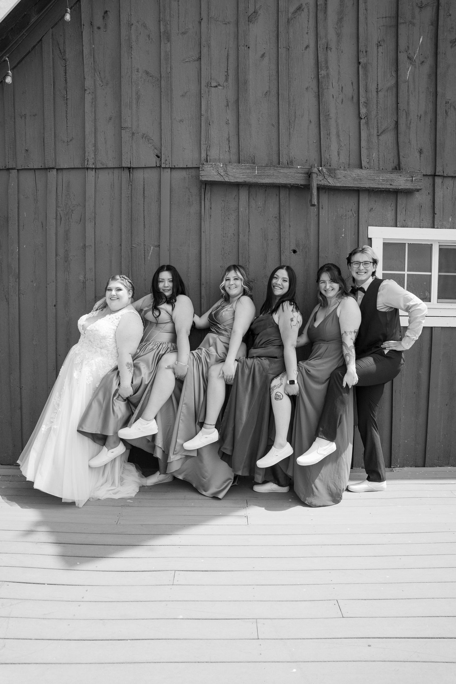 Wedding party members in gray dresses peek playfully around a rustic barn wall in black and white.