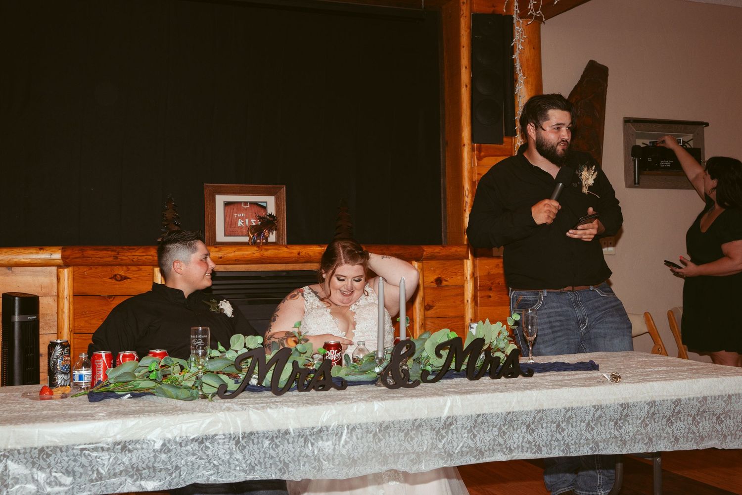 Wedding reception guests seated at long tables inside a rustic wooden venue with warm lighting.
