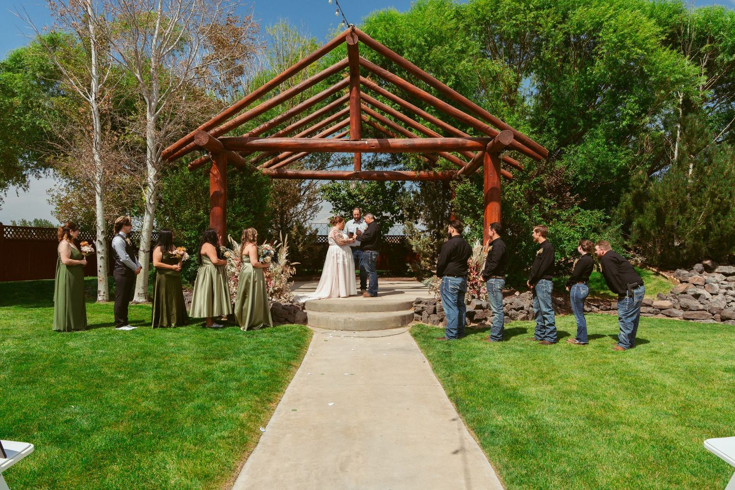 Wedding ceremony taking place under a rustic wooden pavilion on a lush green lawn with guests watching.