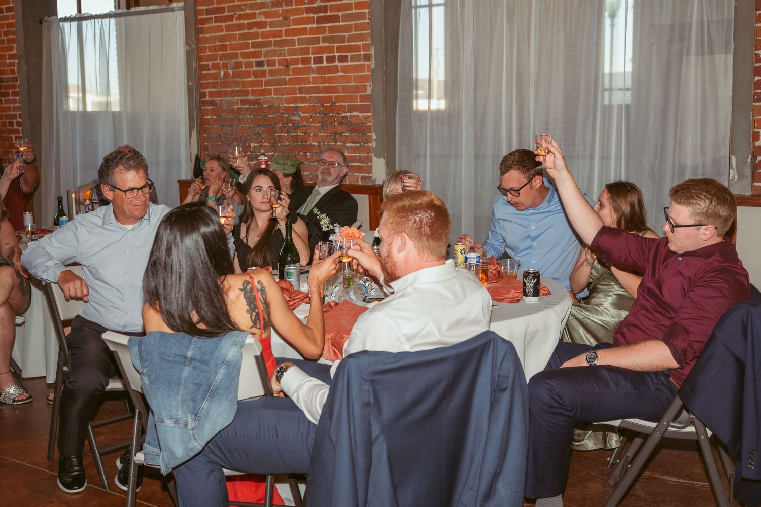 Wedding guests raise their drinks in celebration at tables inside an industrial-style brick venue.