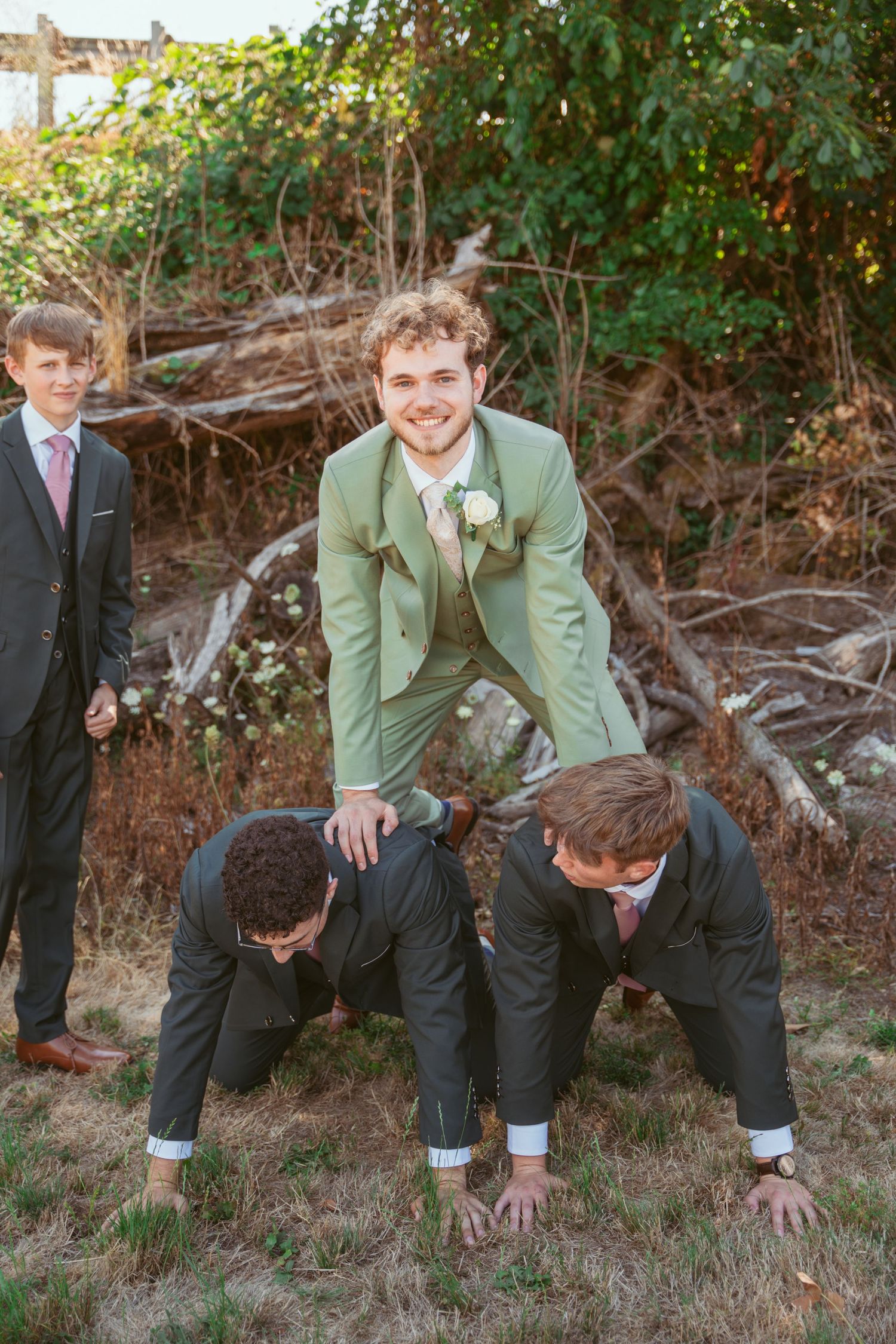 Groomsmen in sage green suits perform playful push-ups on grassy ground during wedding photo session.