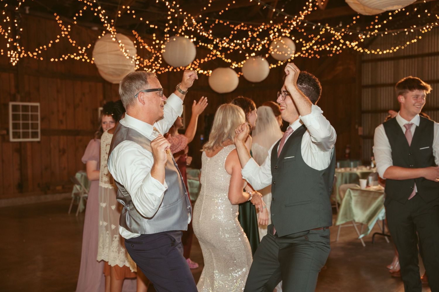 Guests celebrate on the dance floor under romantic string lights and paper lanterns at a barn wedding.