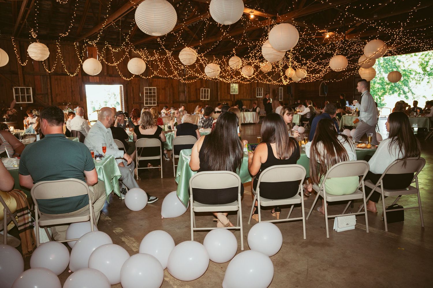 Wedding guests seated in rows of chairs with white balloons decorating a rustic barn venue.