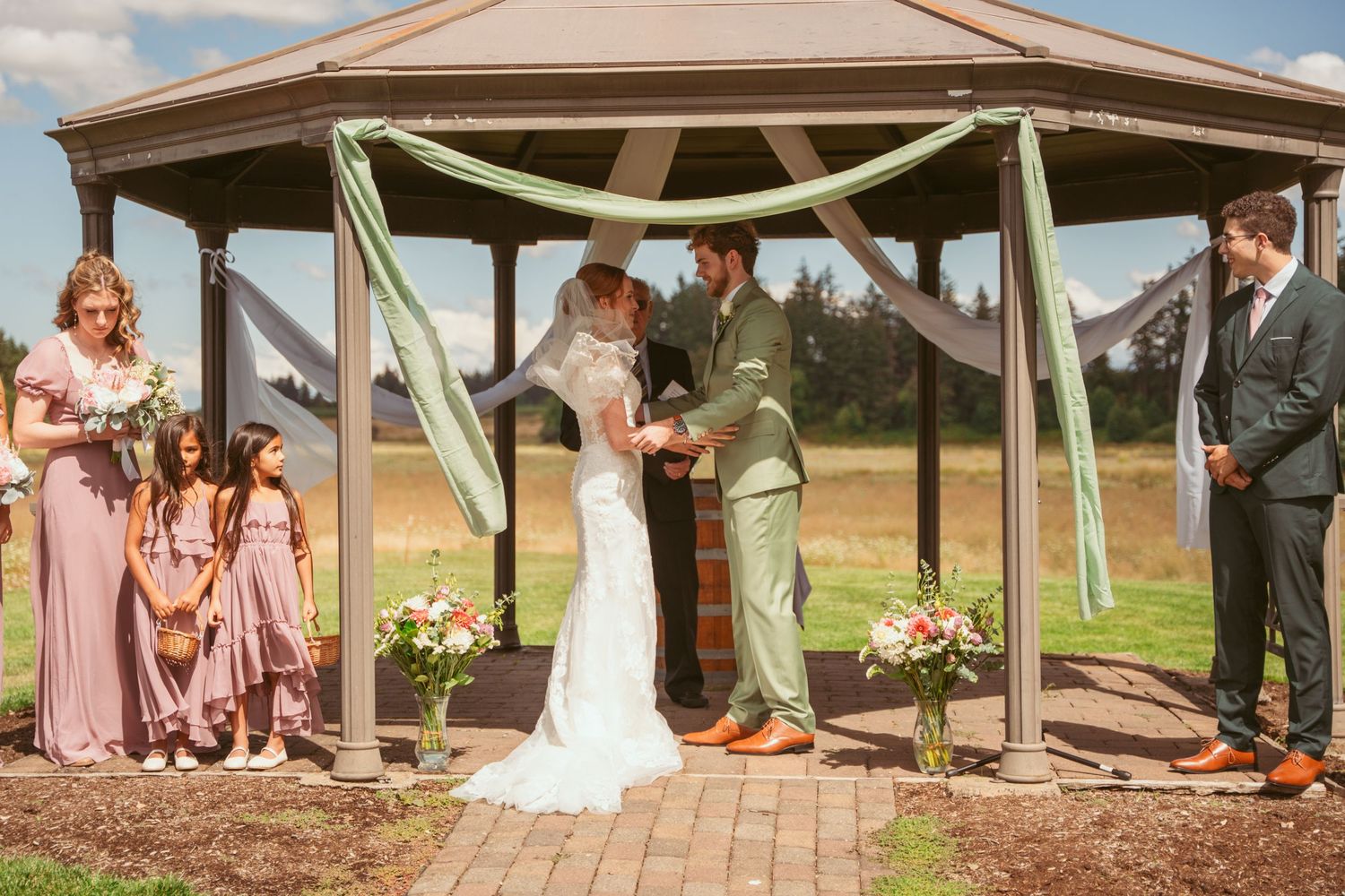 Wedding ceremony in progress under a covered pavilion with pink and green color scheme.