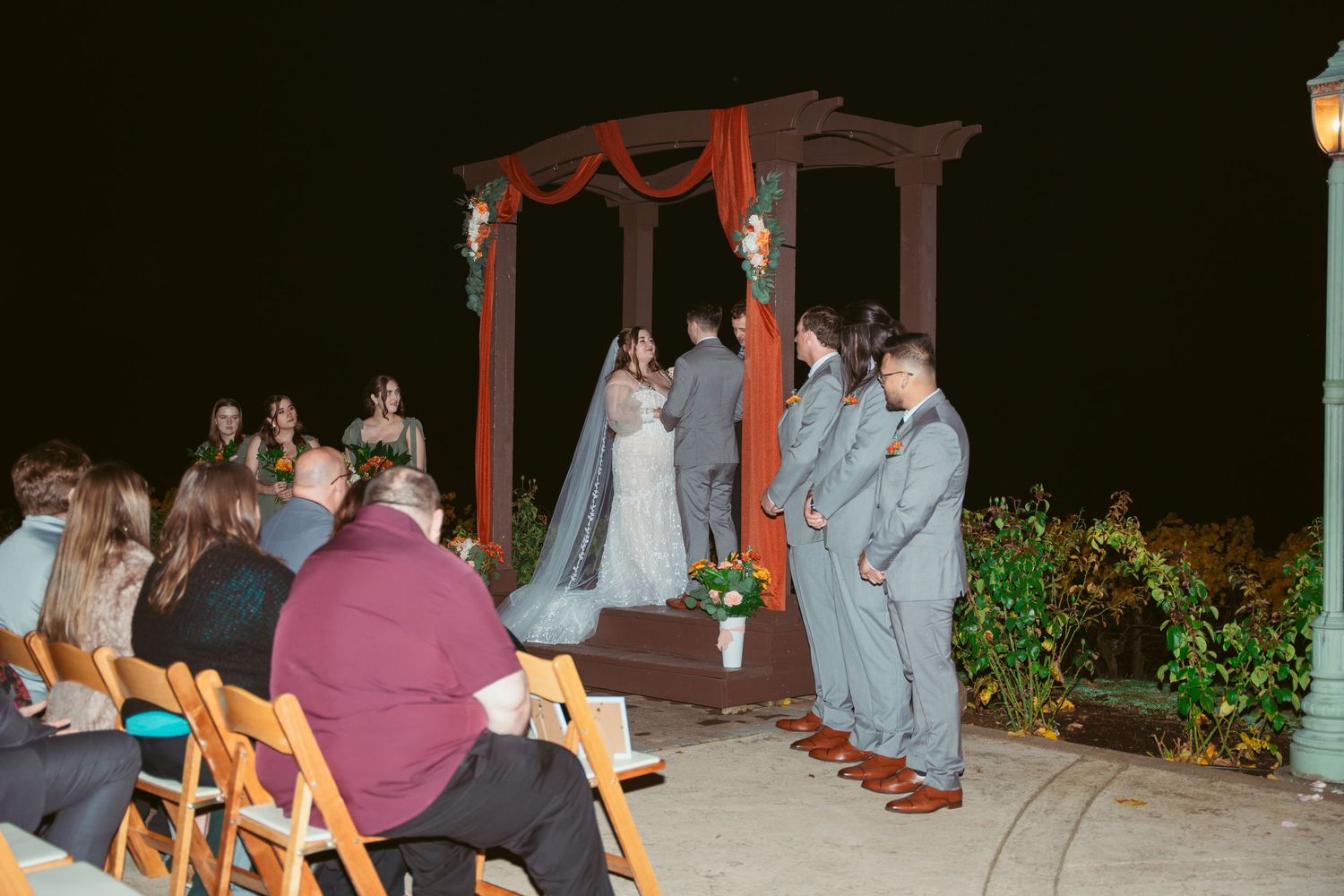 Wedding ceremony under a red wooden arch with guests seated on folding chairs at night.