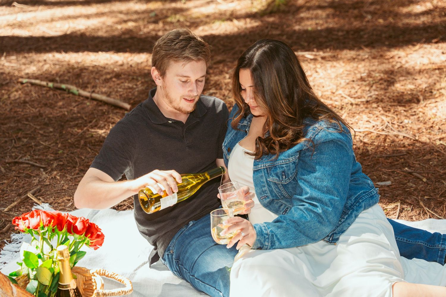 A romantic picnic setup in a forest with a wicker basket, wine bottles, and red roses on a white blanket.