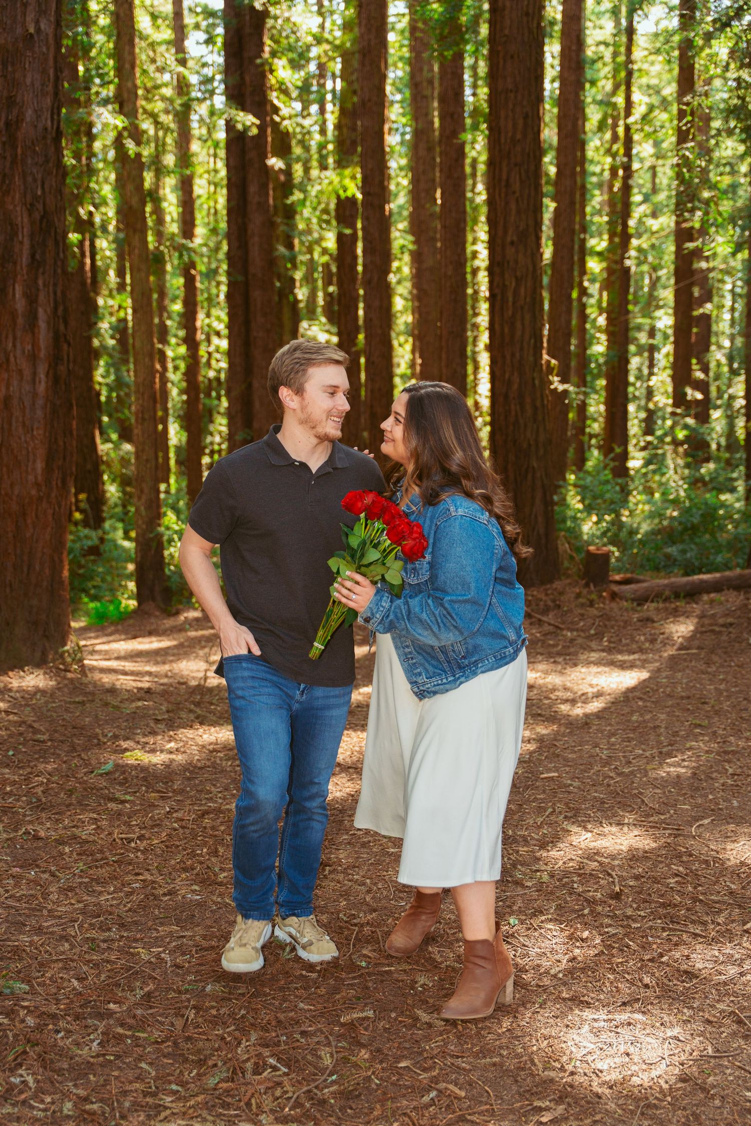 A couple shares a tender moment while walking among towering redwood trees on a forest path.