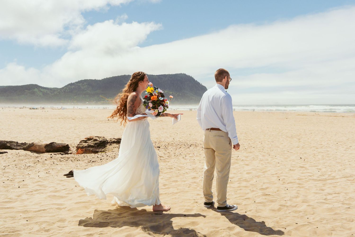 Couple walking along a scenic beach with mountains and blue sky in the background.