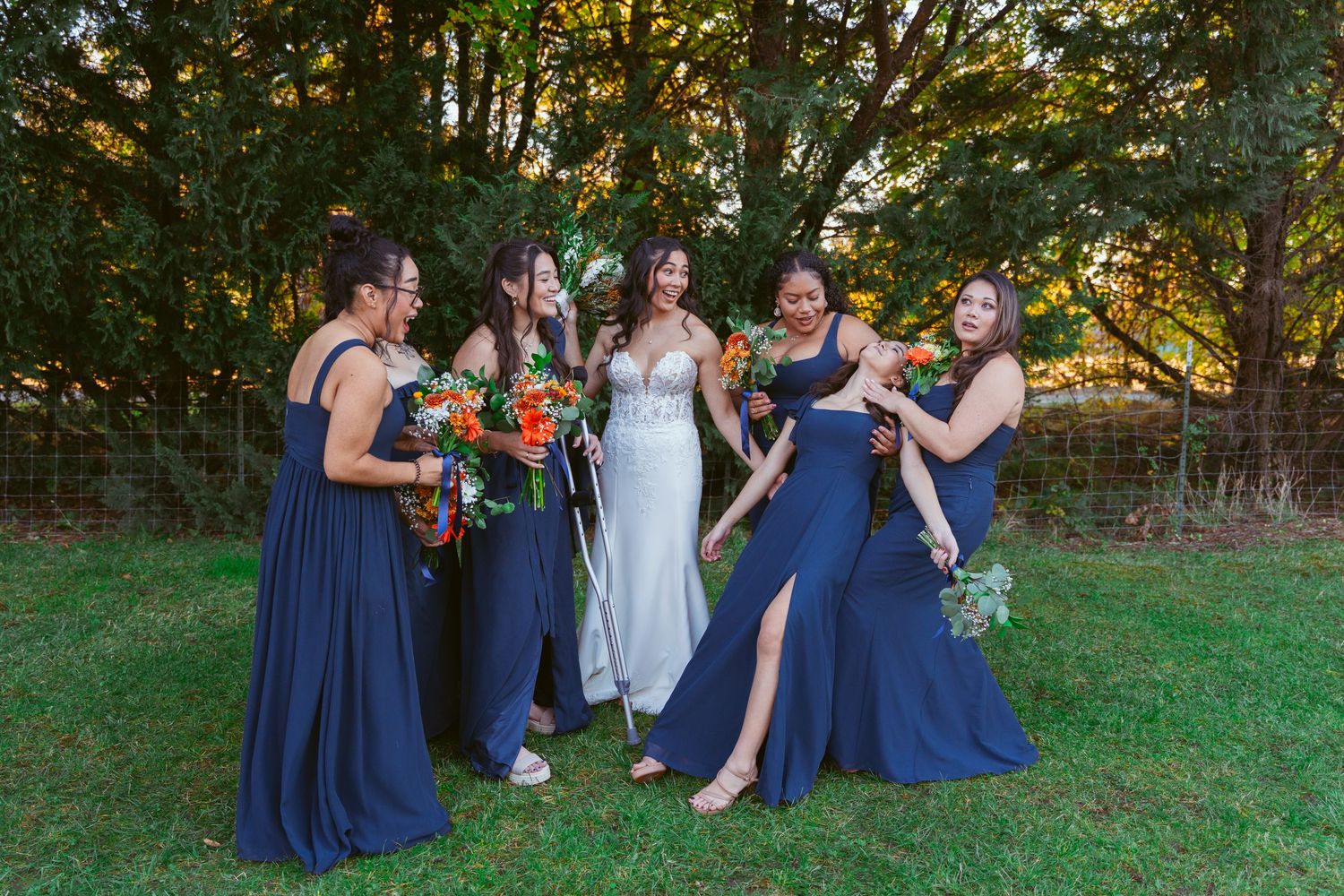 Wedding party in navy and white poses for casual photos on green lawn with orange bouquets.