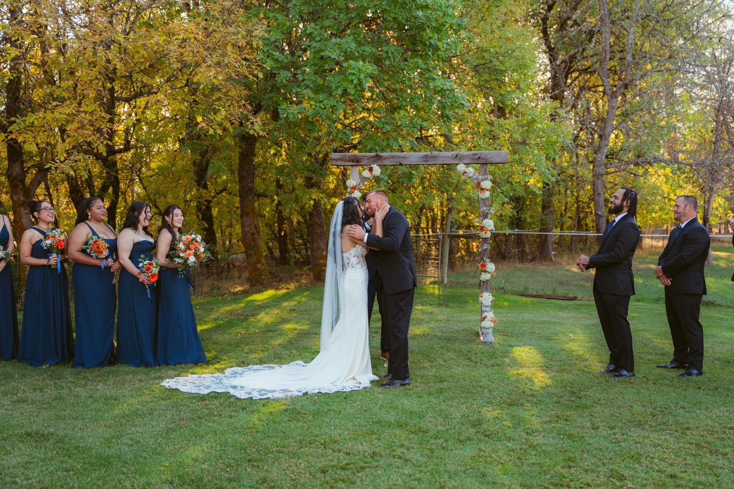 Fall wedding ceremony under an arch with attendants in navy blue dresses against autumn foliage.