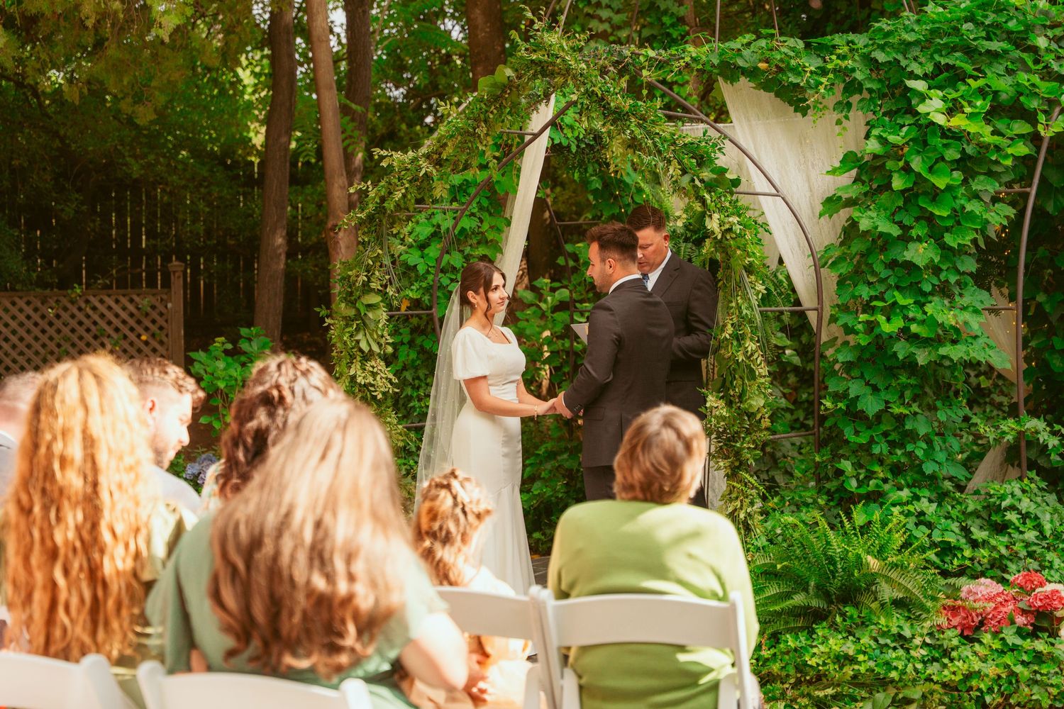 Wedding guests seated in white chairs watch an outdoor ceremony in a lush garden setting.