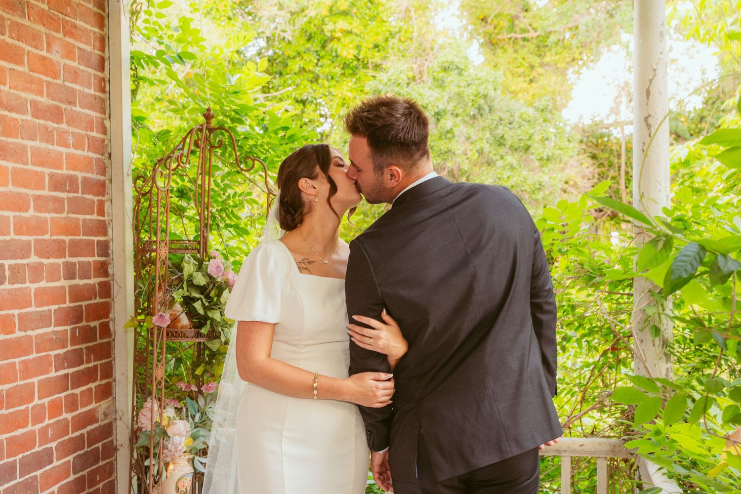 A wedding couple embraces on a brick porch with natural greenery overhead.