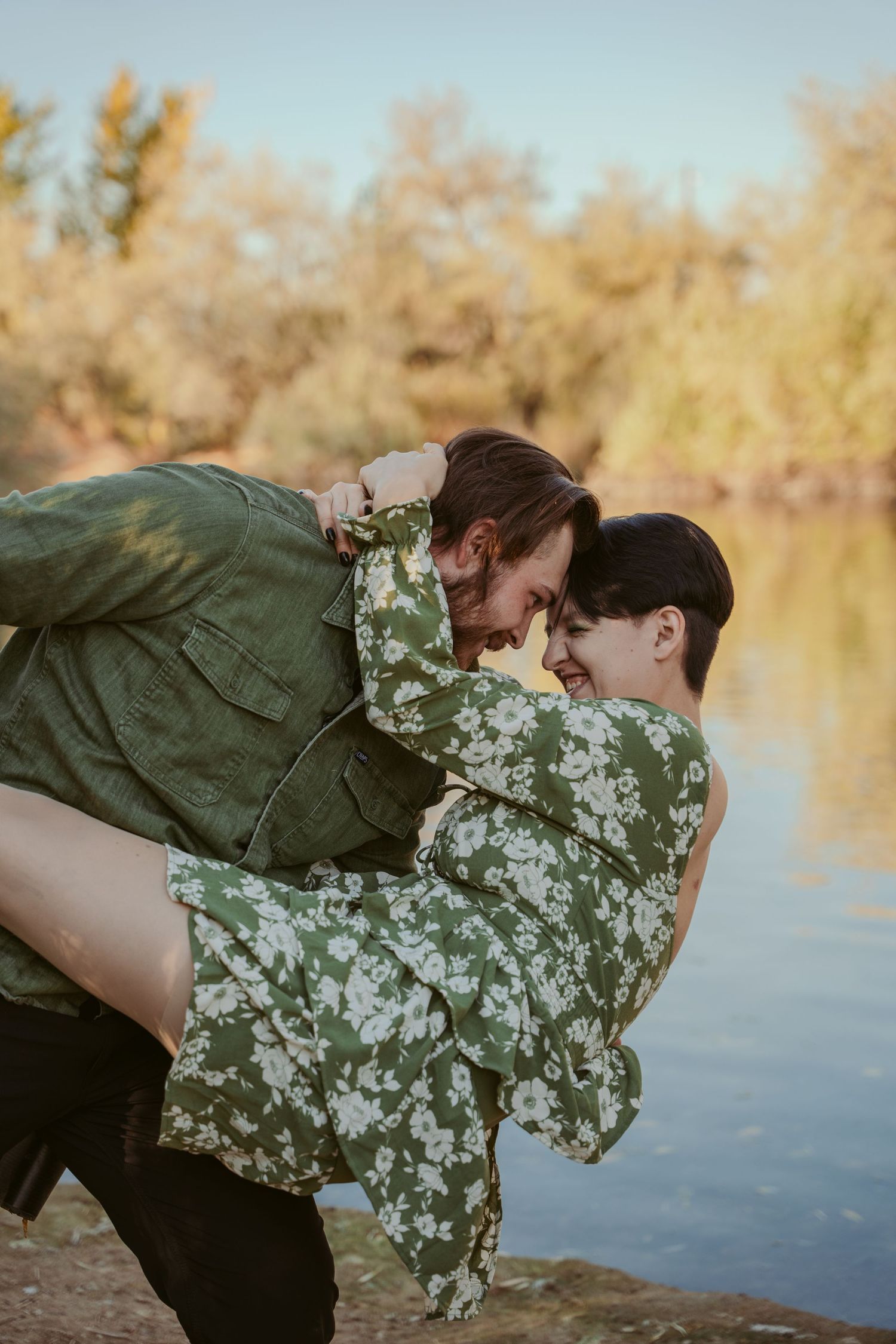 Couple in green attire poses romantically by a serene lake at sunset.