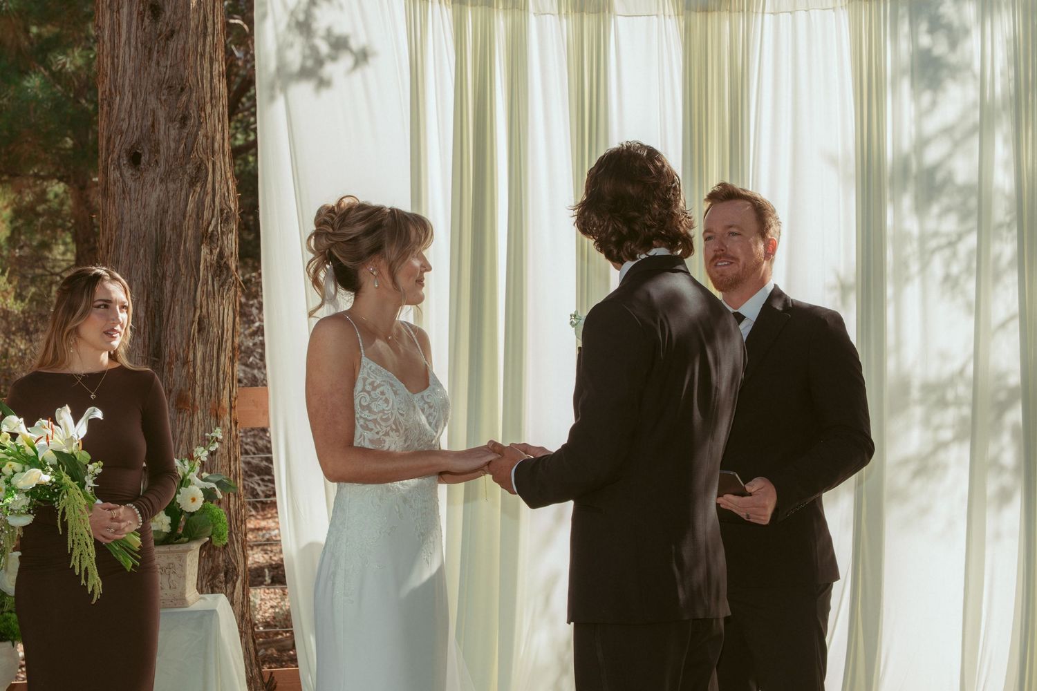 Wedding ceremony in progress under tall trees with white fabric backdrop and natural lighting.