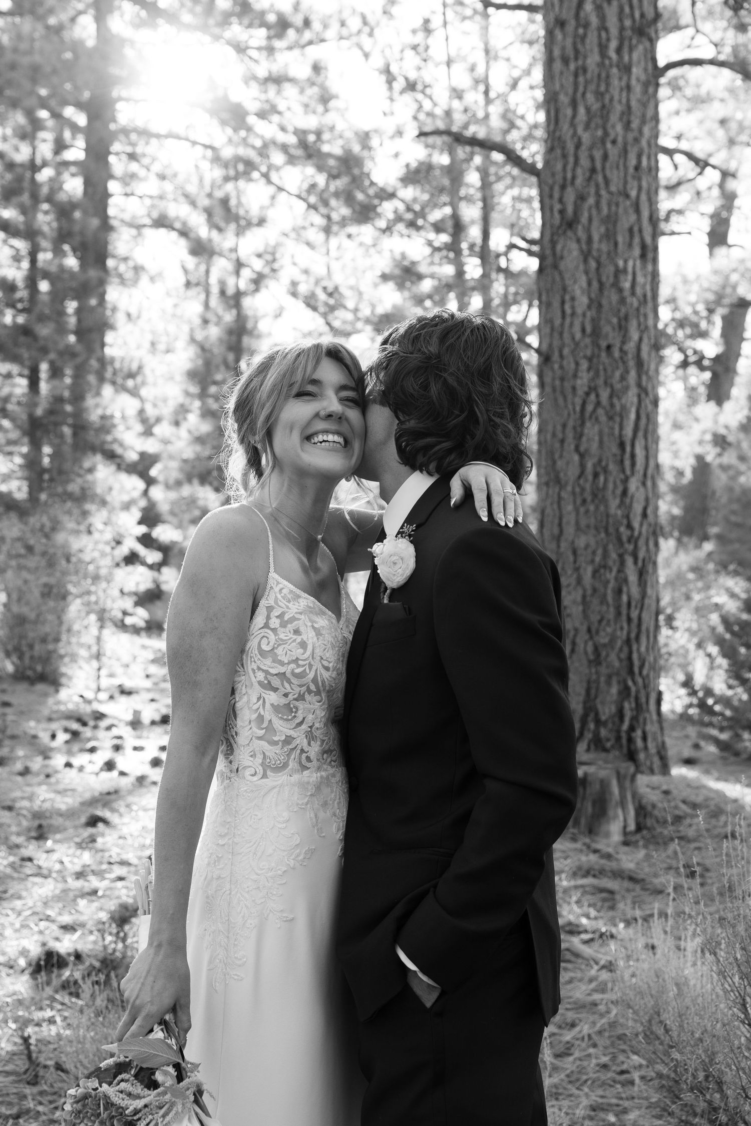 A couple shares a joyful moment together among pine trees in black and white photography.