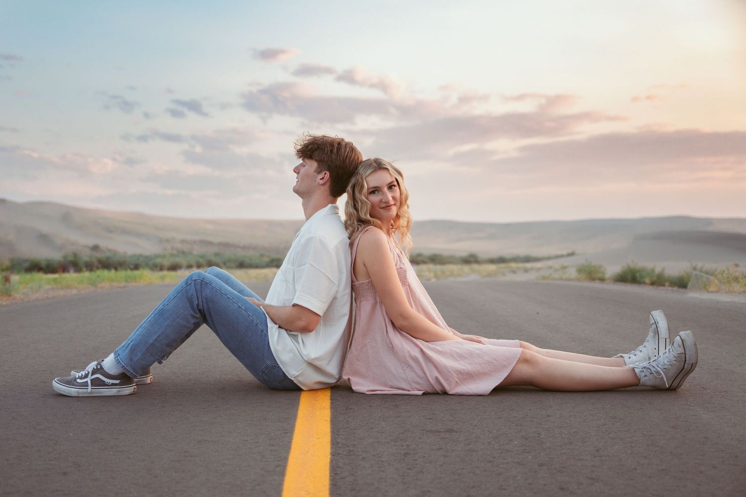 A couple sits back-to-back on a yellow road line during sunset.
